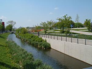 A man in a red shirt overlooks a stream of water flowing beside the pedestrian bridge where he is standing on a bright sunny day.