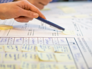 Zoomed in view of someone preparing a report. Someone is reaching across the papers with a pen.