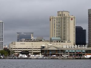 Skyline of the city of Norfolk, VA along the waterfront. There are a multitude of tall buildings and hotels and a shorter, wide building with the name "WATERSIDE" prominently displayed.