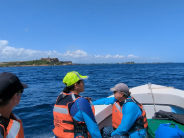 Three people in orange life vests on a small boar in bright blue water on a clear sunny day