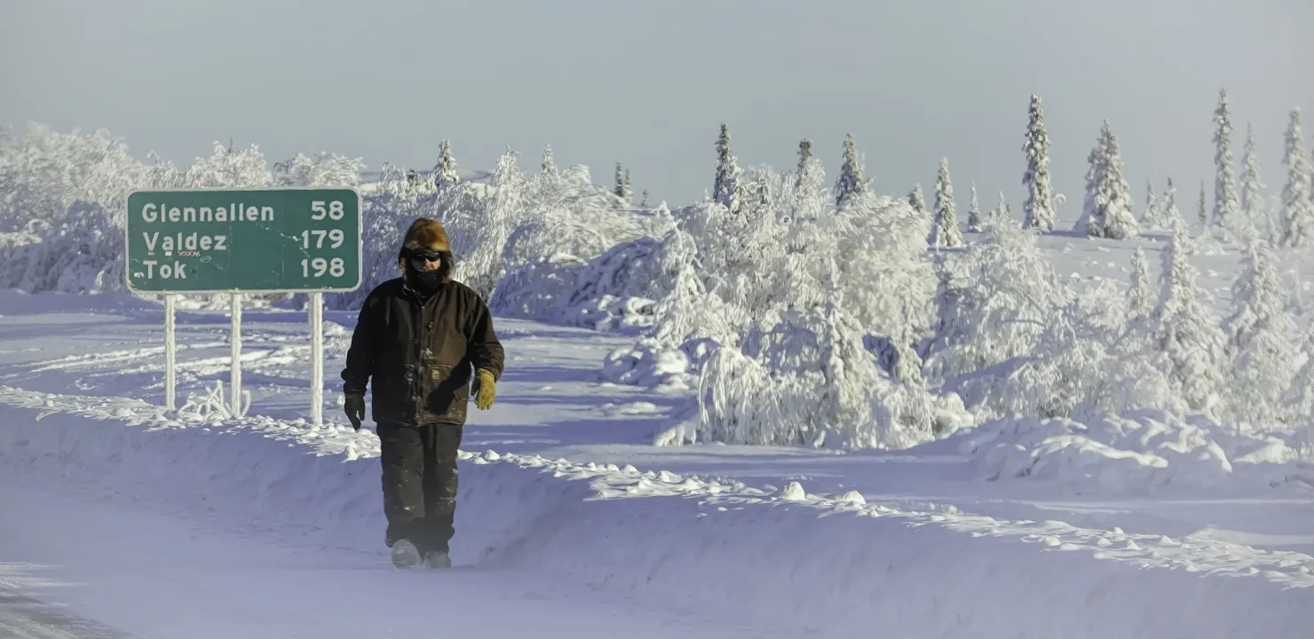 Someone walking toward the camera in the snow in front of a highway sign