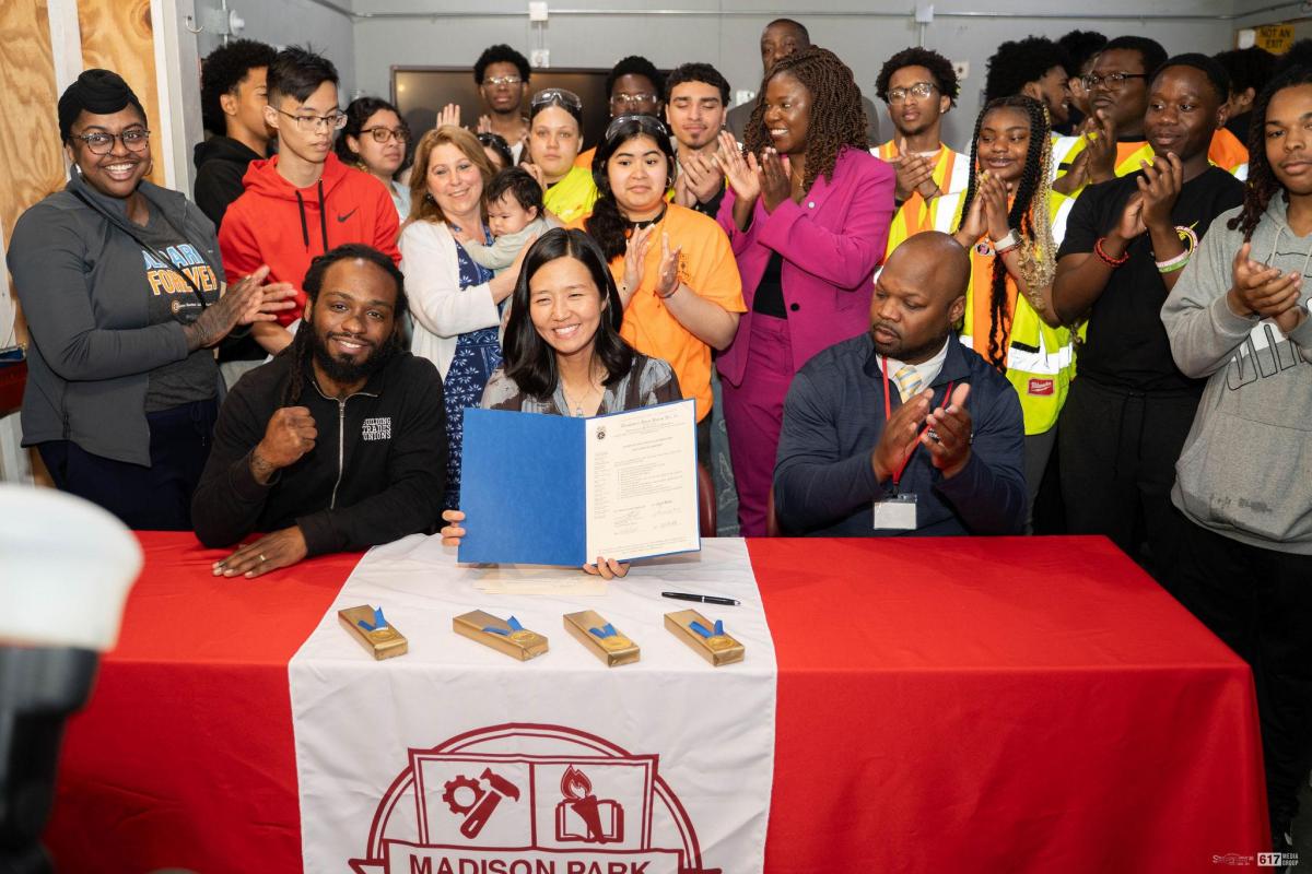 A group of people gathered around a table celebrating as a woman holds up a signed document, smiling. The table is draped with a red cloth featuring the "Madison Park" logo. Some attendees are clapping and cheering in the background.