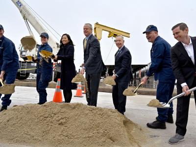 A group of people in work uniforms and suits are participating in a groundbreaking ceremony, each holding a shovel with sand. They are standing around a pile of sand with construction equipment visible in the background.