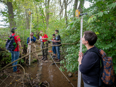 Several students and an instructor conduct stream surveying in a dense, wooded forest.