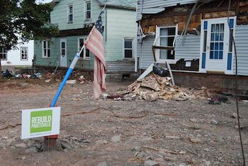 Two homes affected by a storm with structural damage. An American flag stands in the foreground behind a sign reading "Rebuild Prattsville"