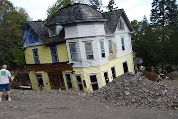 A man stands beside a damaged multicolored house with a pile of debris in front, surrounded by trees.