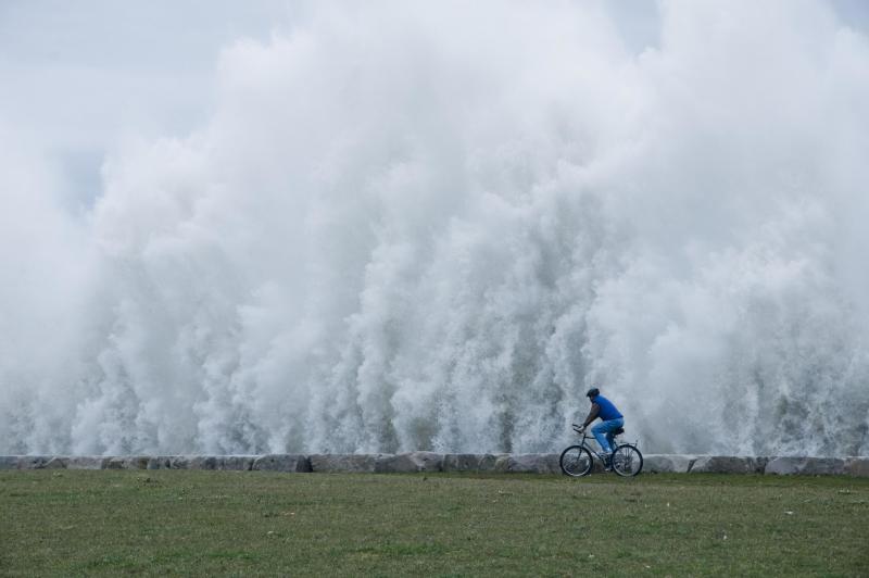 A man riding his bicycle beside an apparent wall of water as winds kick up 20-foot waves along the shores of Lake Michigan.
