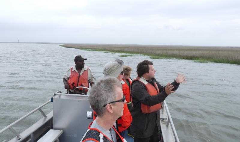 A group of five people in red life jackets look off the side of a boat to the coastal wetland. The man in the front is passionately speaking with his hands stretched forward.