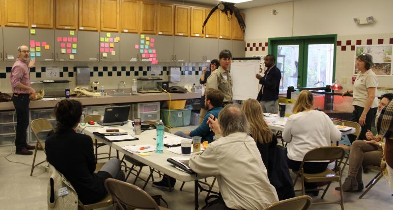 Members of the partnership gather in a classroom and their attention is to the front of the class where an individual points toward various colored sticky notes placed on the cabinets.