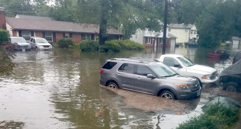 A flooded suburban street lined with homes and various automobiles submerged to their wheel wells in food waters.