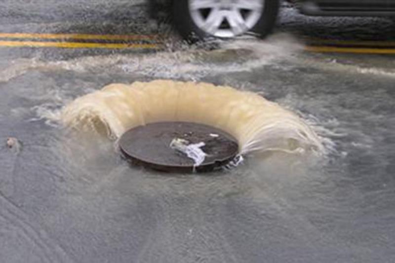 A flooded section of a street and water spraying out from a manhole cover in the middle of the street.