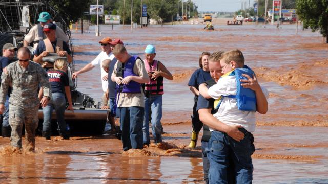 A firefighter carries a young child wearing a life jacket to safety, up to her ankles in muddy floodwater. 