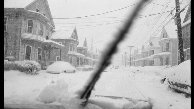 Houses covered in heavy snow on Lawrence Street, MA seen through the snowy windshield of a car