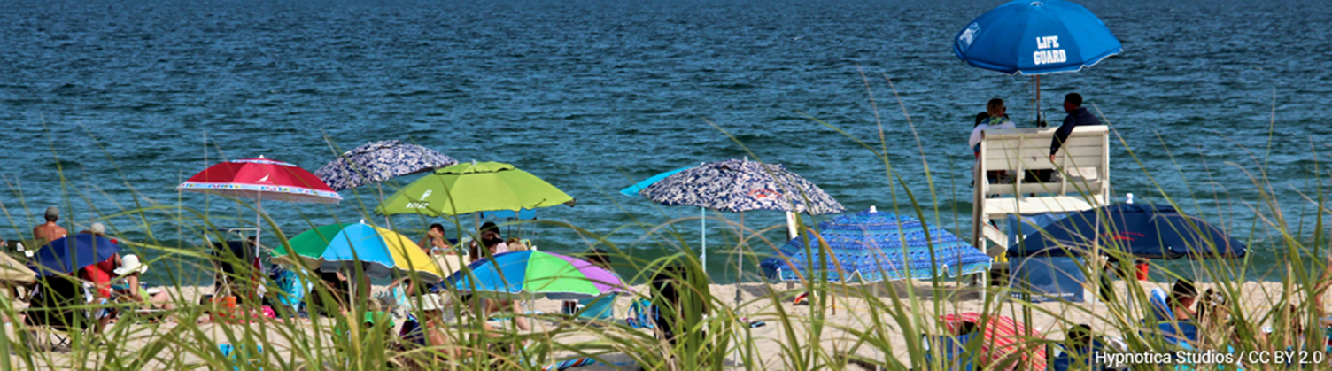 A view from the sand dunes of a group of people sitting at the beach. The bright colorful umbrellas stand out in contrast to the deep blue ocean that takes over the top half of the photo. Two lifeguards sit on a tall lifeguard chair. 